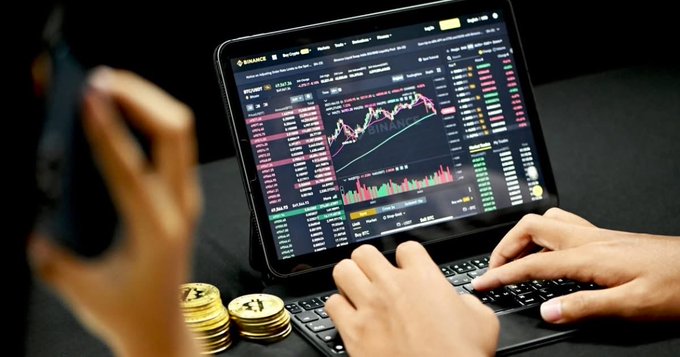 A person holds a phone while typing on a tablet displaying a Binance cryptocurrency trading chart with candlesticks and BTC/USDT data, with stacks of gold Bitcoin tokens on the desk.