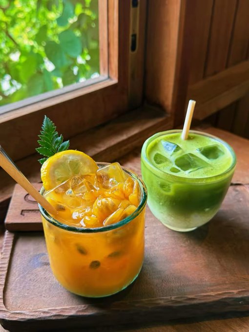 Two refreshing beverages—one orange passion fruit drink with a lemon slice and mint, and one green matcha drink—served with bamboo straws on a wooden tray beside a window.