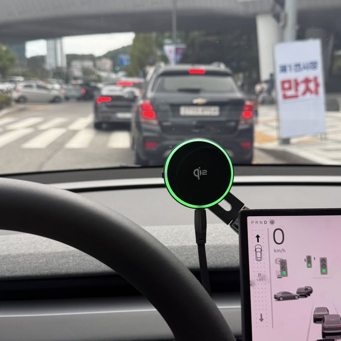 A Tesla dashboard displays a Qi2 wireless charger with its signature green ring mounted near the steering wheel, alongside a touchscreen showing "PRND" and "0 km/h" as the car idles at a red light behind a Chevrolet. The post's humor plays on "Tesla-ram" sounding like "Tesla-rang," suggesting the innovative Qi2 charging experience makes the driver feel like they're "rang"-ing with delight.