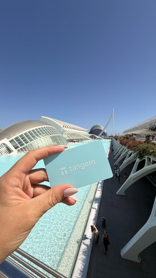 A hand holds up a mint-green "tangem" card against a stunning backdrop of futuristic white architecture and a reflecting pool under a clear blue sky, capturing a memorable sightseeing moment.