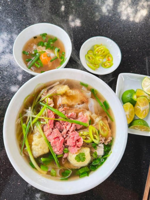 A bowl of Vietnamese noodle soup, likely pho, is served alongside a small bowl of egg drop soup, sliced green chilies, and a plate of lime wedges.