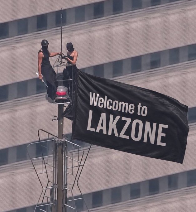 Two people stand atop a tall pole affixing a black "Welcome to LAKZONE" banner, aligning with the post's announcement that invites for the Lakzone event are being sent out and encouraging followers to check their DMs from @lakzonevn.