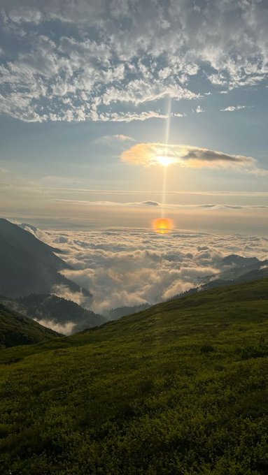 Breathtaking landscape photograph showing a sunset above a sea of clouds in a mountain valley, with the sun creating a sun pillar and casting golden light over the cloud layer.