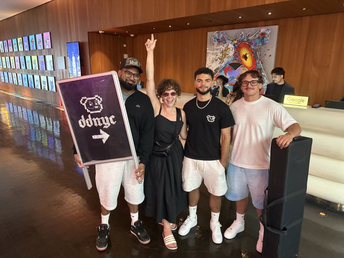A group of four people posing together indoors, with one person holding a directional sign that reads 'oddnyc' with an arrow pointing right.