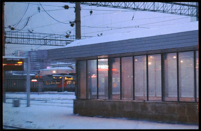 A snowy winter scene at a train station showing an illuminated waiting shelter with large windows, a commuter train in the background, and overhead power lines crossing the dusk sky.