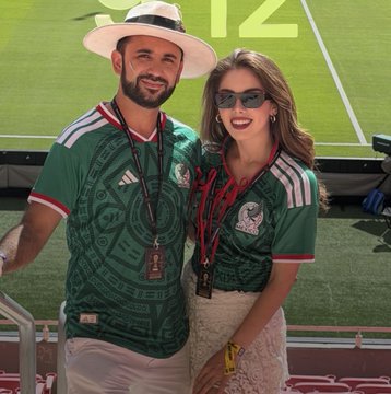 A smiling man wearing a sombrero and a green Mexico national team jersey stands next to a woman in sunglasses at a stadium.