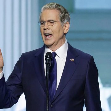 A man in a dark blue suit with an American flag pin speaking at a podium with a microphone.