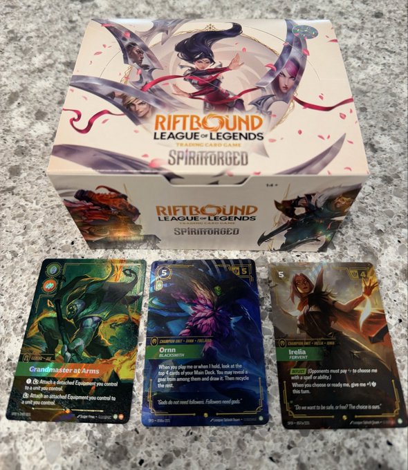 A Riftbound: League of Legends Trading Card Game "Spiritforged" booster box is displayed alongside three cards pulled from it, including a holographic "Grandmaster at Arms," a standard "Ornn - Blacksmith," and an alternate-art "Irelia - Perfexpert" (likely foil/overnumbered), celebrating the collector's excitement over their first overnumbered pull.
