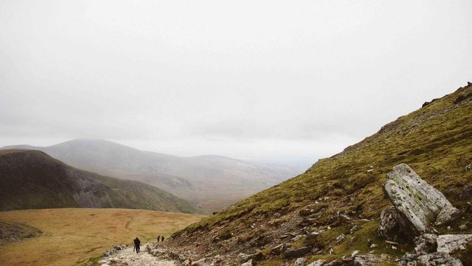 Hikers traverse a misty, rugged mountainside path beneath an overcast sky, with a moss-covered boulder dominating the foreground. The image accompanies a post about the fading hype around NFTs, though it has no visual connection to crypto topics—likely attached as a stock landscape photo.