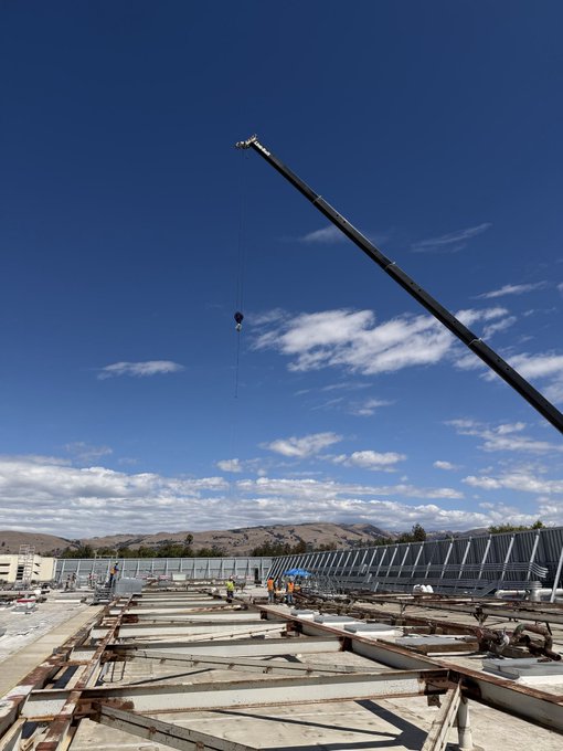 A construction site with steel beams and concrete under a bright blue sky with scattered clouds, where a large crane is lifting materials with workers visible in the distance.