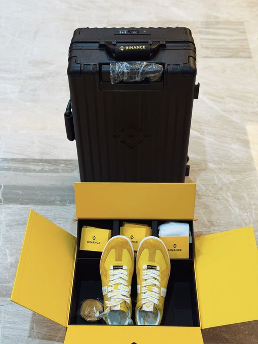 A black 'BINANCE' branded suitcase stands behind an open yellow box containing yellow sneakers and Binance-branded accessories, all displayed on a marble floor.