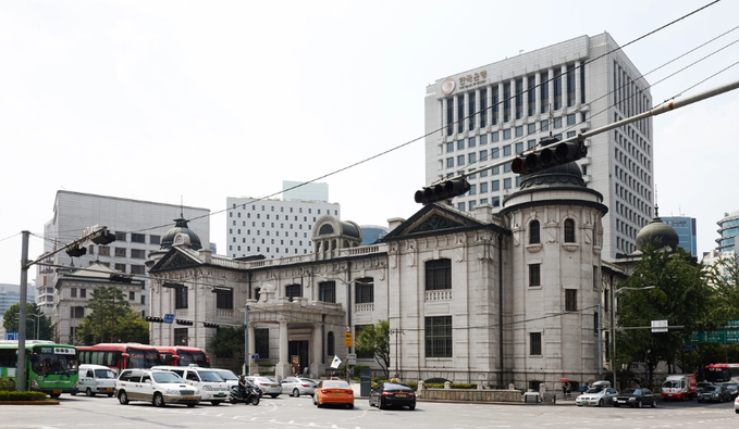 A historic European-style building stands in the foreground with modern skyscrapers behind it, including a Bank of Korea building visible in the background, while city traffic moves through the intersection in Seoul. The image relates to the breaking news about South Korea's new cryptocurrency reporting requirements, as the Bank of Korea building in the background represents the central bank that will be receiving exchange records of crypto withdrawals to personal wallets.