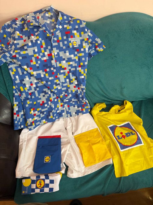 A Lidl-branded children's outfit set laid out on a green couch, including a blue pixel-patterned shirt, white shorts with blue and yellow pockets, and a yellow Lidl tote bag.