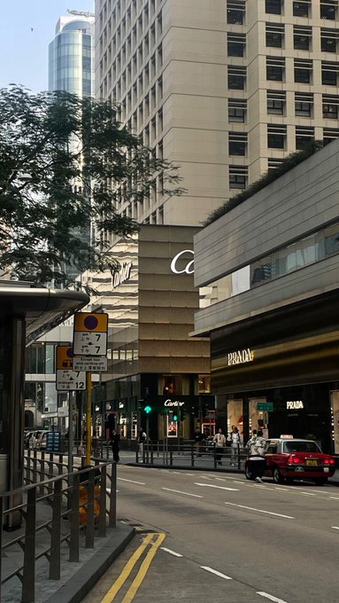 A street view in Hong Kong showing the facade of a Cartier store and a Prada store, with a red taxi driving past a traffic light that shows a 'No Left Turn' sign.