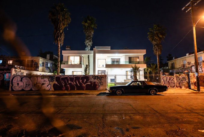 A classic lowrider parked in front of the modern "Hood Mansion" at night, surrounded by graffiti-covered walls reading "GODS LIKE" and "KING" under palm trees—visually echoing the post's $HOODMAN meme token's nod to barrio street culture.