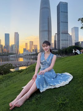 A young woman in a blue floral dress sits smiling on a grassy hill overlooking a modern city skyline at sunset. The image pairs with the post's bullish $HYPE prediction to $100, using her cheerful "good morning fam" pose against an upscale urban backdrop to visually hype optimism around the token.