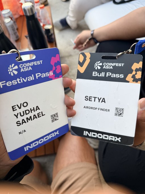Two Coinfest Asia passes are being held up by hand—Evo Yudha Samael's blue "Festival Pass" and Setya's black "Bull Pass" for Airdrop Finder—both sponsored by INDODAX, capturing a meet-up moment at the event.