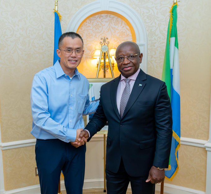Two men in formal attire—one in a light blue button-down shirt and the other in a dark suit with a patterned tie—shake hands while smiling, standing between two flags in an elegant interior setting.