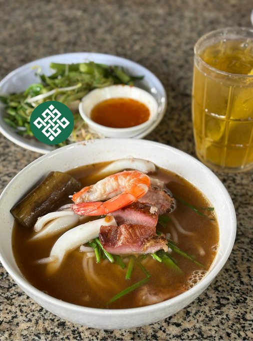 A steaming bowl of Vietnamese soup features wide rice noodles topped with pink shrimp, sliced beef, and green herbs in a rich brown broth, accompanied by a side plate of fresh greens, a small dish of dipping sauce, and a tall glass of iced tea. The image pairs with the post "Good food. Good mood. @ritualnet" to showcase a satisfying, mood-boosting meal.