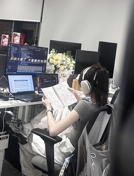 A person wearing headphones sitting at a desk with multiple monitors displaying code, reading a printed document.
