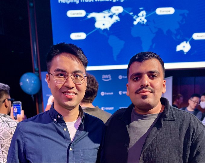 Two men pose together at what appears to be a Trust Wallet side event, with a world map display behind them highlighting connections between locations like Canada, Europe, USA, and Australia. The image relates to the caption by capturing the international, community-focused atmosphere described, with the two smiling attendees representing the diverse, global crowd that gathered at the event.