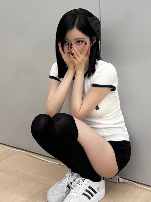 A young woman crouches against a gray wall wearing a white t-shirt with black trim, black thigh-high socks, white Adidas sneakers, and a black hair bow, with her hands covering her face.