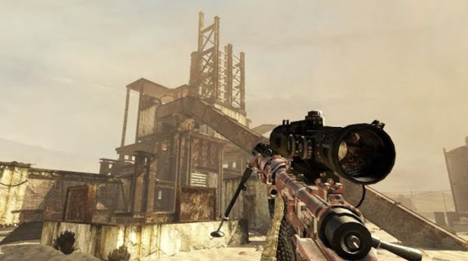 First-person view from a video game showing a sniper rifle aimed at an industrial structure in a desert setting.