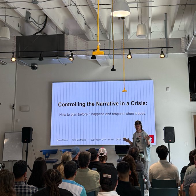 In the image, a woman stands behind a screen with a presentation slide titled "Controlling the Narrative in a Crisis" displayed at the front of the room. The audience consists of people sitting in chairs around her, attentively listening to her speech or presentation. A microphone is visible on the right side of the image, likely being used by the woman speaking into it.