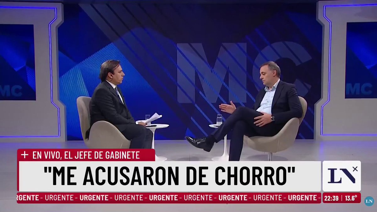 The image features a man sitting in a chair, talking with another person who appears to be wearing a suit. The background of the scene includes a blue wall with white text that reads "MCC" and "En Vivo Jeffé de Gabinee". There is also an article about Bitcoin on the screen, which seems to be related to the conversation between the two individuals.