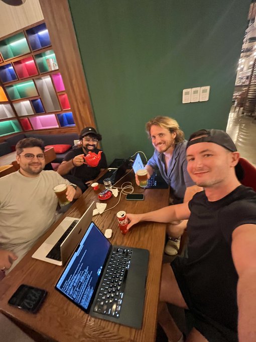 A group of four men gathered around a table in what appears to be a cafe, with laptops, coffee, and Coca-Cola cans visible, one man pouring from a red teapot.