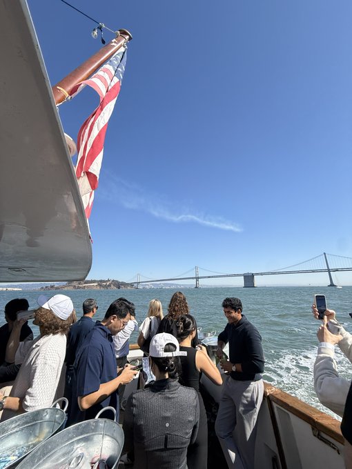 Passengers stand on the deck of a boat with an American flag, traveling on the water toward a large suspension bridge in the distance under a clear blue sky.