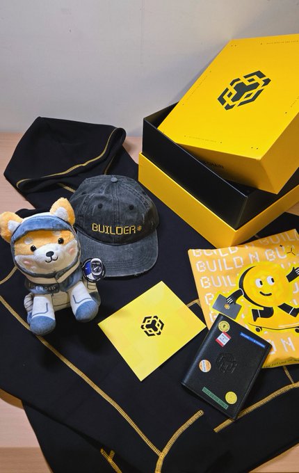 The image features a black box with yellow writing, which appears to be a gift or present for someone named @BNBCHAINZH. The box contains various items such as a stuffed animal, a hat, and some cards. There is also a wallet inside the box. In addition to these objects, there are two other small items in the image: a teddy bear and a card with a picture of a yellow flower on it.