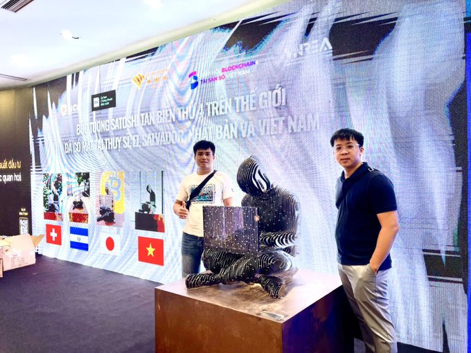 Two young men pose alongside a metallic sculpture of "Satoshi Nakamoto" at what appears to be the SATOSHI ICON exhibition in Ho Chi Minh City, with the displayed text reading "SỰ KIỆN TRƯNG BÀY SATOSHI ICON THỨ 4 TRÊN THẾ GIỚI" (the 4th Satoshi Icon exhibition in the world), featuring flags of Switzerland, Honduras, Japan, and Vietnam.