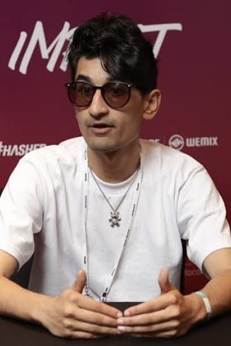 A young man with dark hair and tinted glasses wearing a white t-shirt and a necklace sits at an event with "WEMIX" and "#HASHE" branding visible on the backdrop behind him. The image appears to show FrankDeGods, who just confirmed that similar photos circulating online are doctored images from 3-4 years ago being misused to promote suspicious crypto projects.