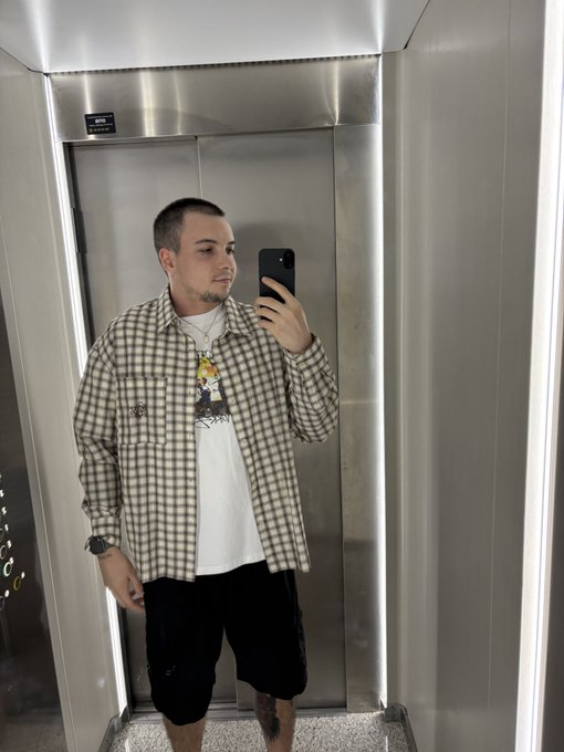 A young man with short hair takes a mirror selfie in an elevator, wearing a brown and white plaid overshirt, a graphic t-shirt, and black shorts.