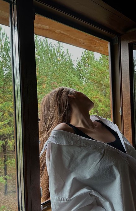 A person with long hair wearing an oversized white shirt over a black top stands by an open wooden window, tilting their head back with their face out of frame, looking out at a forested landscape.