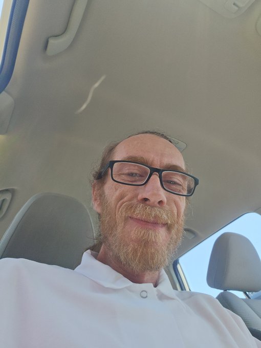 A selfie of a bearded man wearing black-rimmed glasses and a white shirt, sitting inside a car.
