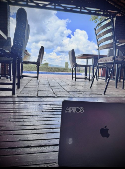 An Apple MacBook laptop sits on a wooden patio table overlooking outdoor seating, a pool, and a cloudy sky.