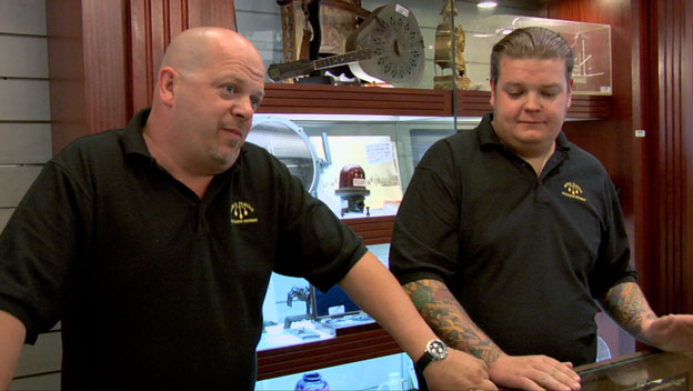 Two men in black polo shirts with 'Gold & Silver Pawn Shop' logos stand inside the shop, with a bald man making a playful expression next to a tattooed coworker.