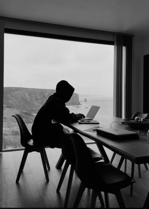 A hooded figure works on a laptop at a modern desk overlooking a coastal cliff view, embodying the remote, luxurious lifestyle suggested by the post's message of financial success through crypto gains—balancing work and leisure in a scenic setting.