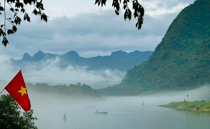 Misty mountain landscape with the Vietnamese flag (red with yellow star) in the foreground, featuring small boats floating on a calm river surrounded by forested peaks.