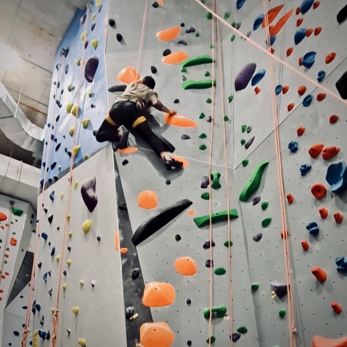 An indoor rock climbing scene showing a person wearing dark clothing with yellow accents scaling a wall covered in colorful climbing holds, secured by ropes.