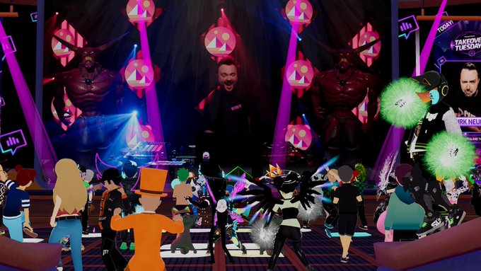 The image shows a lively scene of people dancing and having fun, with some dressed up as characters from video games or movies. The main focus is on a DJ performing for an audience at a party. There are several people standing around, enjoying themselves in various positions within the frame. A person wearing a black shirt can be seen near the center of the image, while another individual stands closer to the right side. In total, there are twelve visible people and three distinct objects: a DJ booth with a microphone on top, a stage with a large screen behind it, and a group of people standing around in front of the stage. The overall atmosphere appears to be energetic and engaging as everyone participates in the party's festivities.