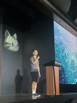 A man speaking at a HashKey podium on stage, with a large screen behind him displaying a cartoon cat logo.