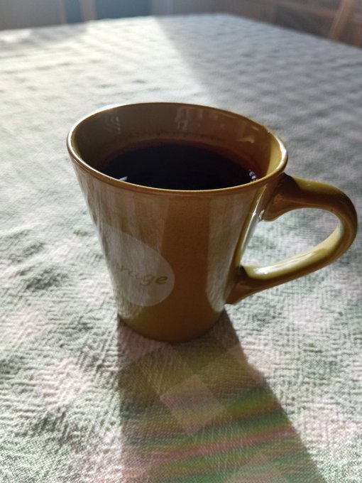 A brown ceramic mug filled with black coffee sits on a textured tablecloth, with sunlight casting a distinct shadow and rainbow prism effect on the cloth.
