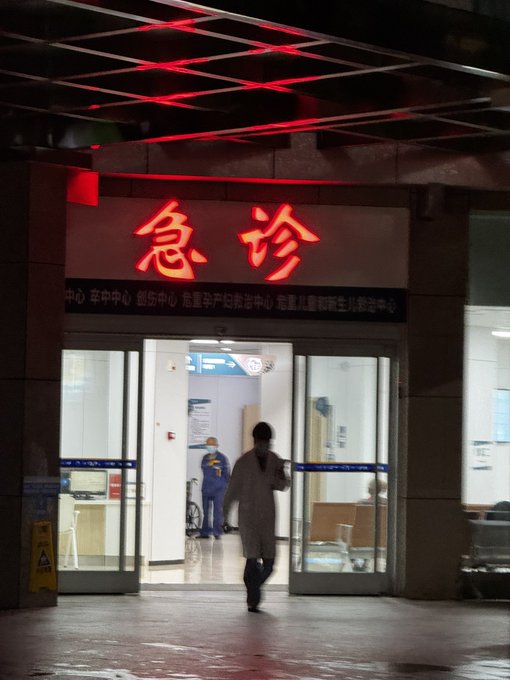 A nighttime photo of a hospital entrance with illuminated red Chinese characters '急诊' (Emergency) above the doors, where a person in dark clothing is walking inside, with medical staff visible in blue scrubs behind the glass.