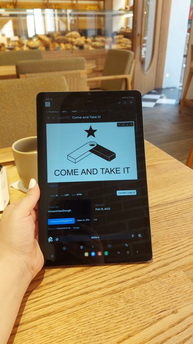 Hand holding a Samsung Galaxy tablet displaying an NFT listing titled 'Come and Take It!' showing a 3D-rendered block design by VmwareLiarv2Dough, with a coffee shop setting blurred in the background.