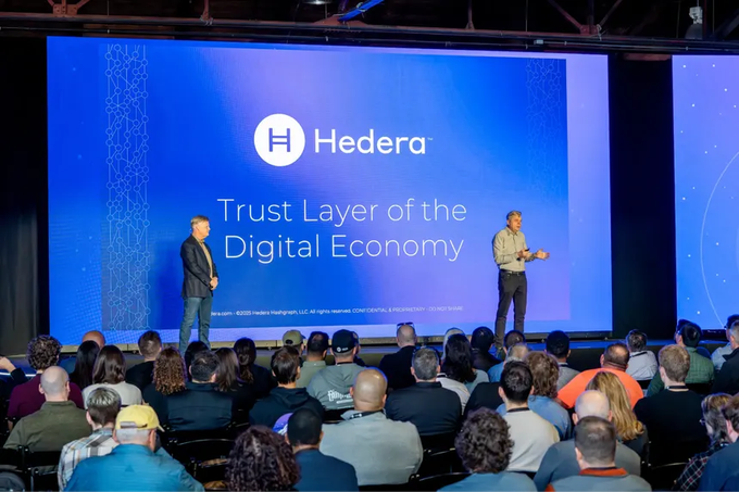 Two presenters stand on stage at a Hedera event in front of a large screen displaying the Hedera logo and the tagline "Trust Layer of the Digital Economy," addressing a seated audience of attendees. The presentation aligns with the post's narrative of Hedera Hashgraph ($HBAR) as a major enterprise blockchain governed by tech giants like Google and IBM.