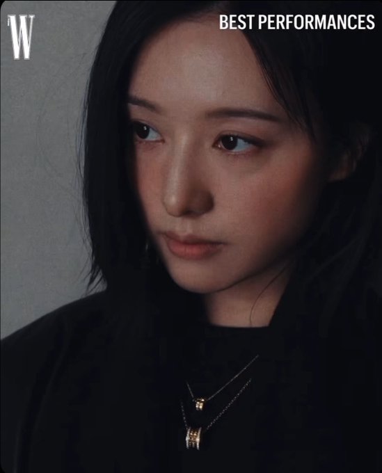 A woman with dark hair and a contemplative expression wears layered gold necklaces against a dark top, while "BEST PERFORMANCES" text appears in the upper right of this W magazine image, ironically highlighting the contrast between polished editorial glamour and the casual gaming reference about spending funds on the Daybreak skin.