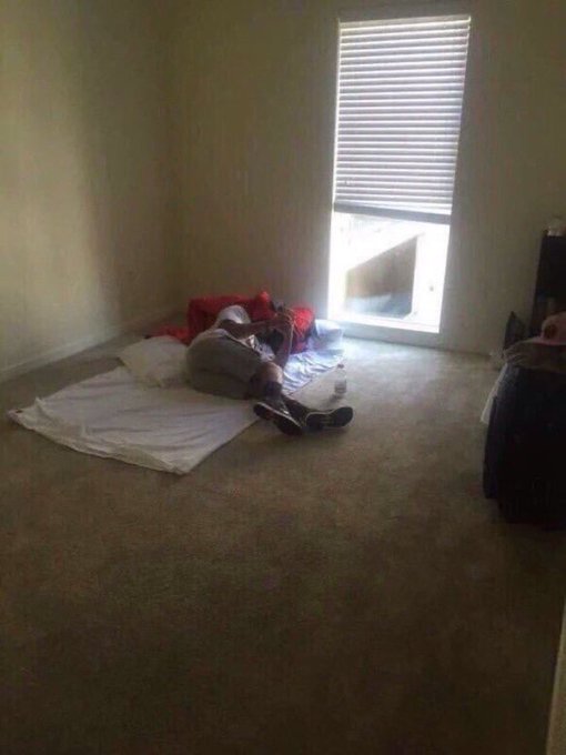 A dimly lit room with carpet shows a person lying on a white blanket and pillow near a glass door with blinds, while another individual sits on the floor against the wall on the right side.